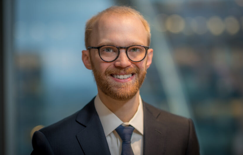 A person with glasses, a beard, and short hair smiles while wearing a dark suit and tie. The background is blurred, featuring a modern office setting.