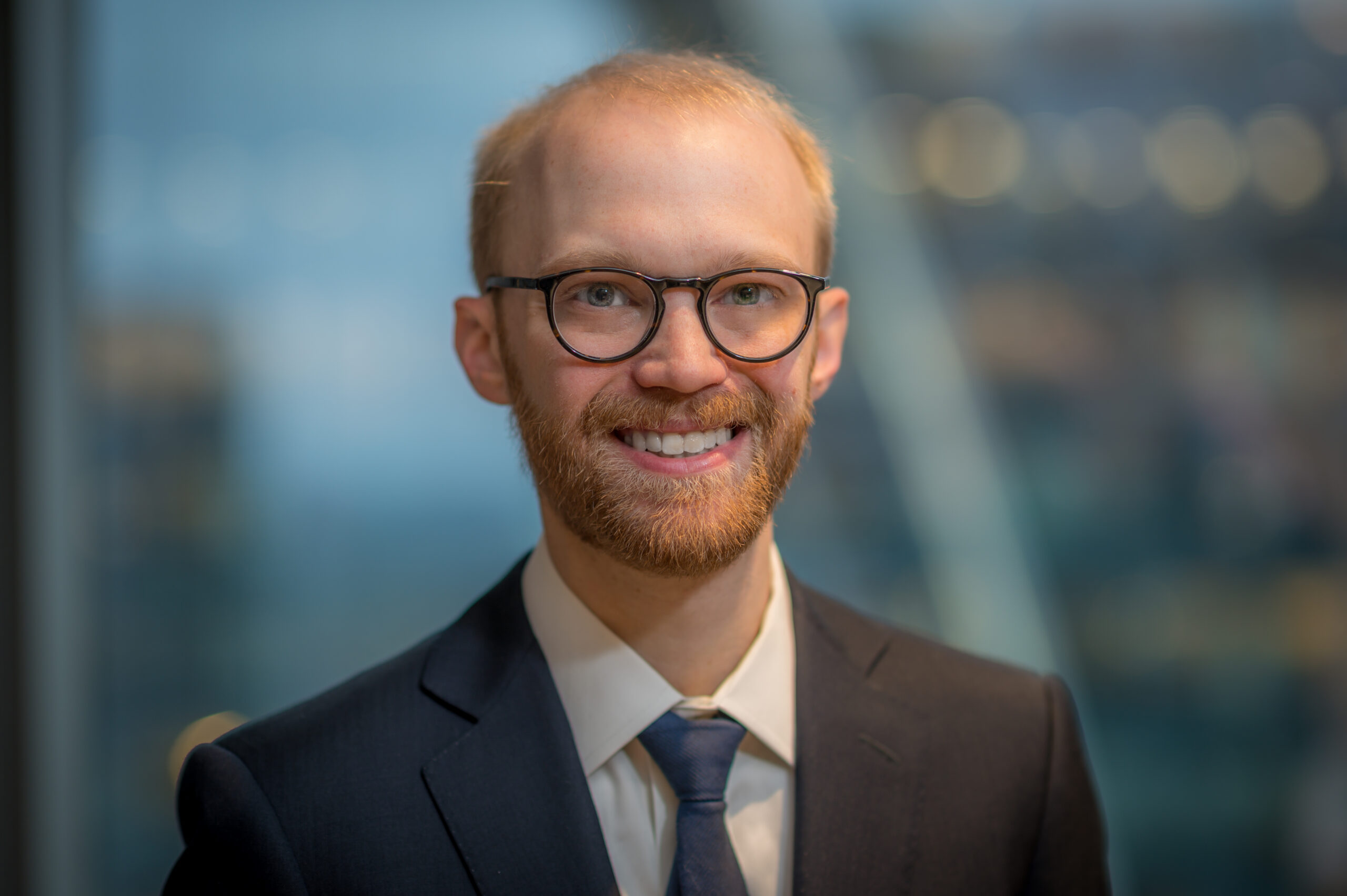 A person with glasses, a beard, and short hair smiles while wearing a dark suit and tie. The background is blurred, featuring a modern office setting.
