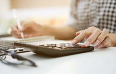 A person wearing a checkered shirt is using a calculator and writing in a notebook on a desk. A laptop and a pair of glasses are also visible in the background.