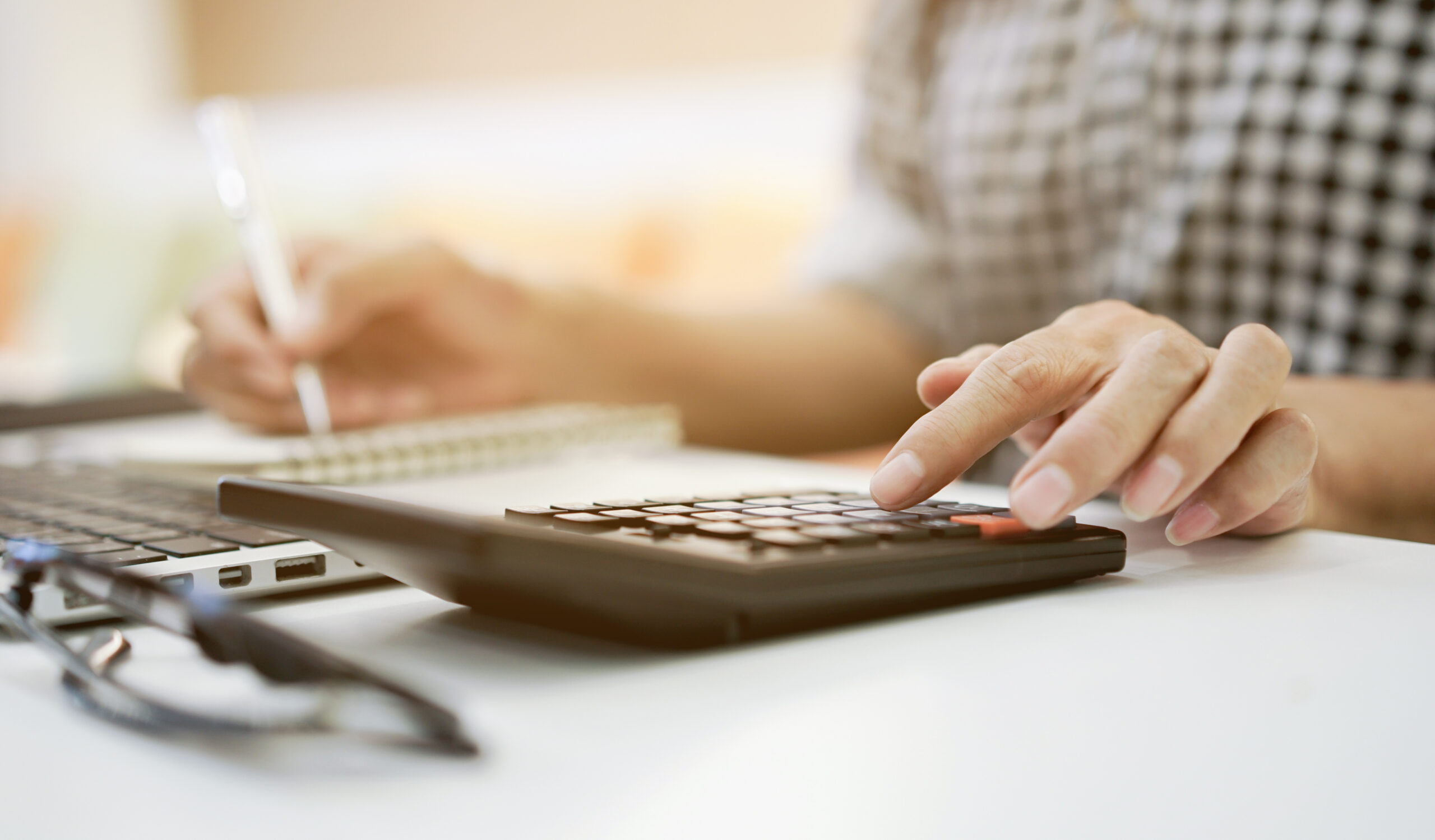 A person wearing a checkered shirt is using a calculator and writing in a notebook on a desk. A laptop and a pair of glasses are also visible in the background.