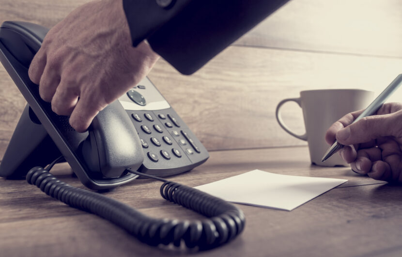 A person picks up a handset from an office phone with one hand while holding a pen over a blank notepad with the other. A coffee cup sits nearby on a wooden desk.