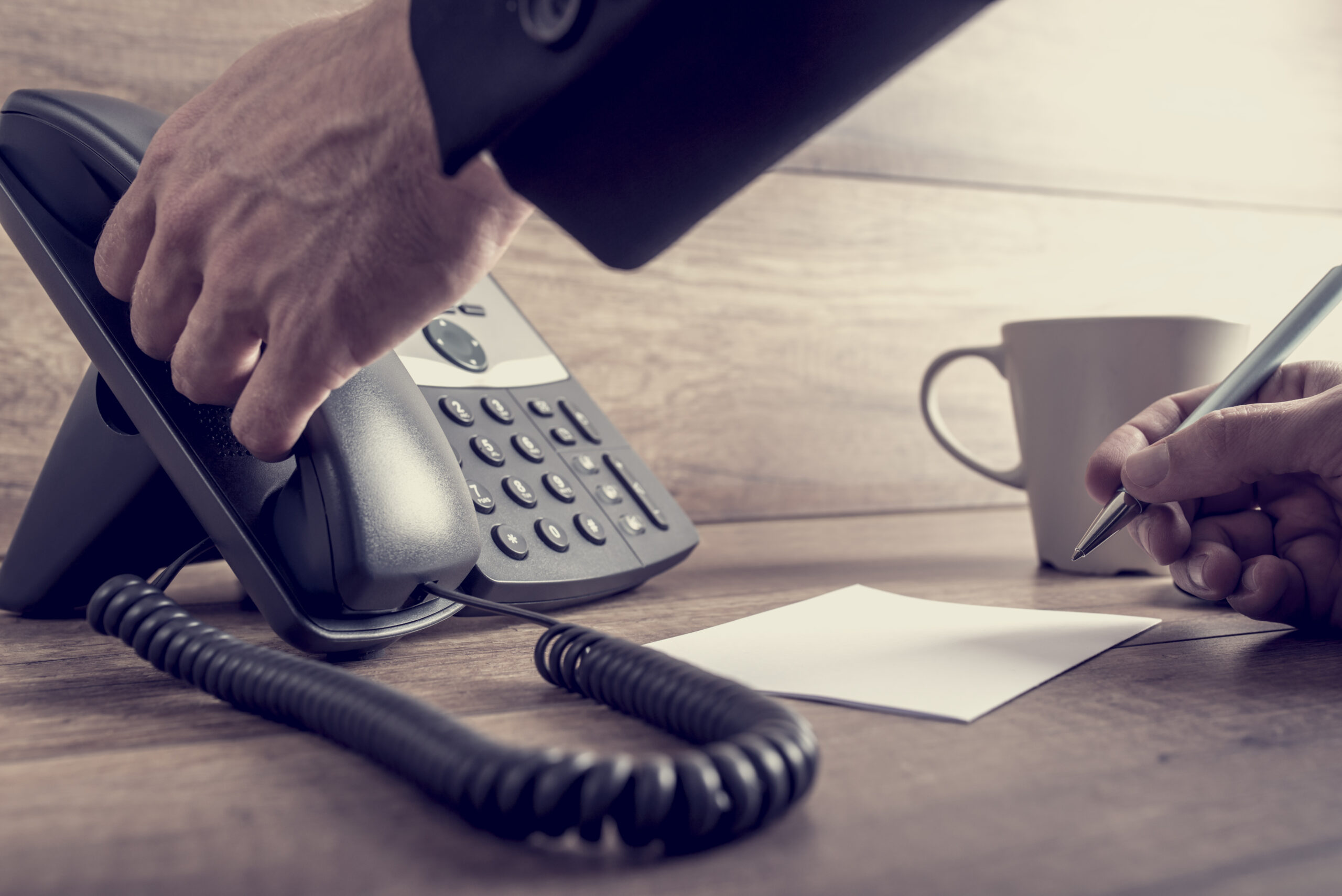 A person picks up a handset from an office phone with one hand while holding a pen over a blank notepad with the other. A coffee cup sits nearby on a wooden desk.