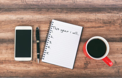 A smartphone, a black pen, an open notebook with This year I will... and a list numbered one to five, and a red cup of coffee are arranged on a wooden surface.