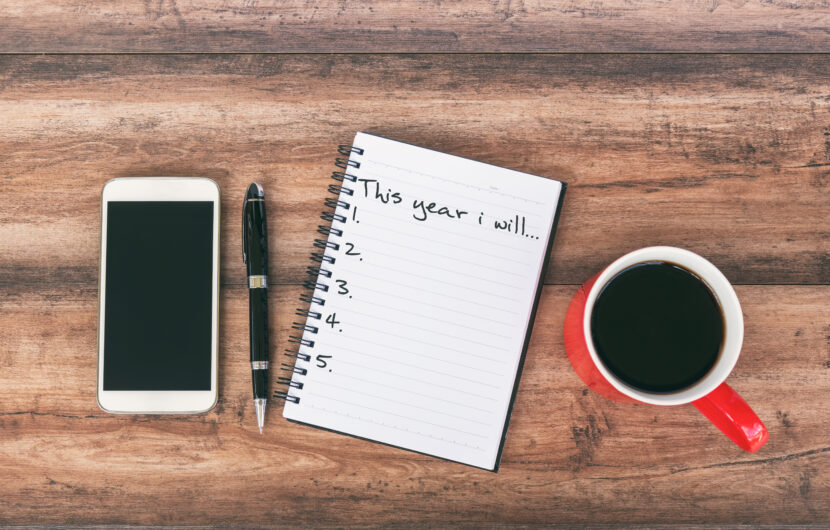 A smartphone, a black pen, an open notebook with This year I will... and a list numbered one to five, and a red cup of coffee are arranged on a wooden surface.