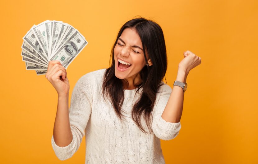 A woman in a white sweater holds a fan of U.S. dollar bills in one hand while pumping her other fist in the air. Her eyes are closed, and she has a joyful expression. The background is solid orange.