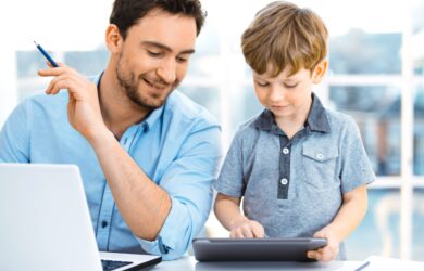 A man smiles and watches a young boy using a tablet. They are seated at a table with a laptop and some papers in a bright room.