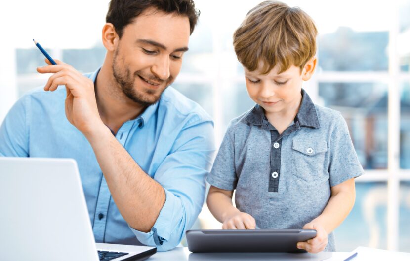 A man smiles and watches a young boy using a tablet. They are seated at a table with a laptop and some papers in a bright room.