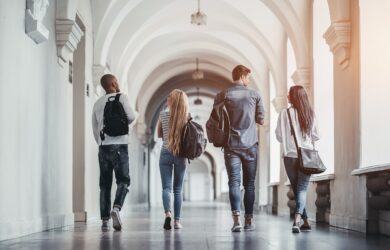Four students walk down a long hallway with high arches and white walls. They carry backpacks and wear casual clothing, heading toward a bright light at the end of the corridor. The image captures their backs as they move forward in unison.