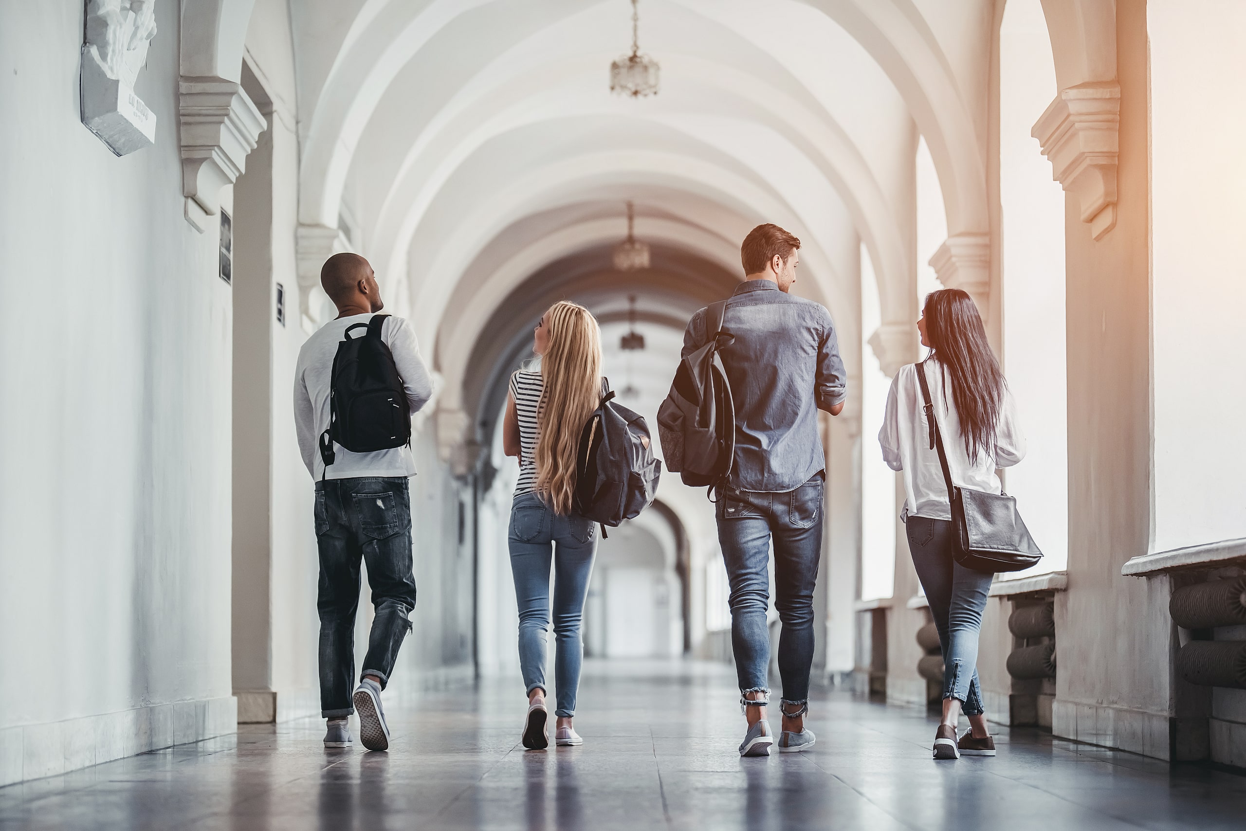 shutterstock_685407808 Four students walk down a long hallway with high arches and white walls. They carry backpacks and wear casual clothing, heading toward a bright light at the end of the corridor. The image captures their backs as they move forward in unison.