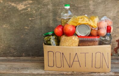 A cardboard box labeled DONATION is filled with various food items, including pasta, canned goods, apples, a bottle of oil, pickles in a jar, and eggs. The box is placed on a wooden surface with a rustic background.