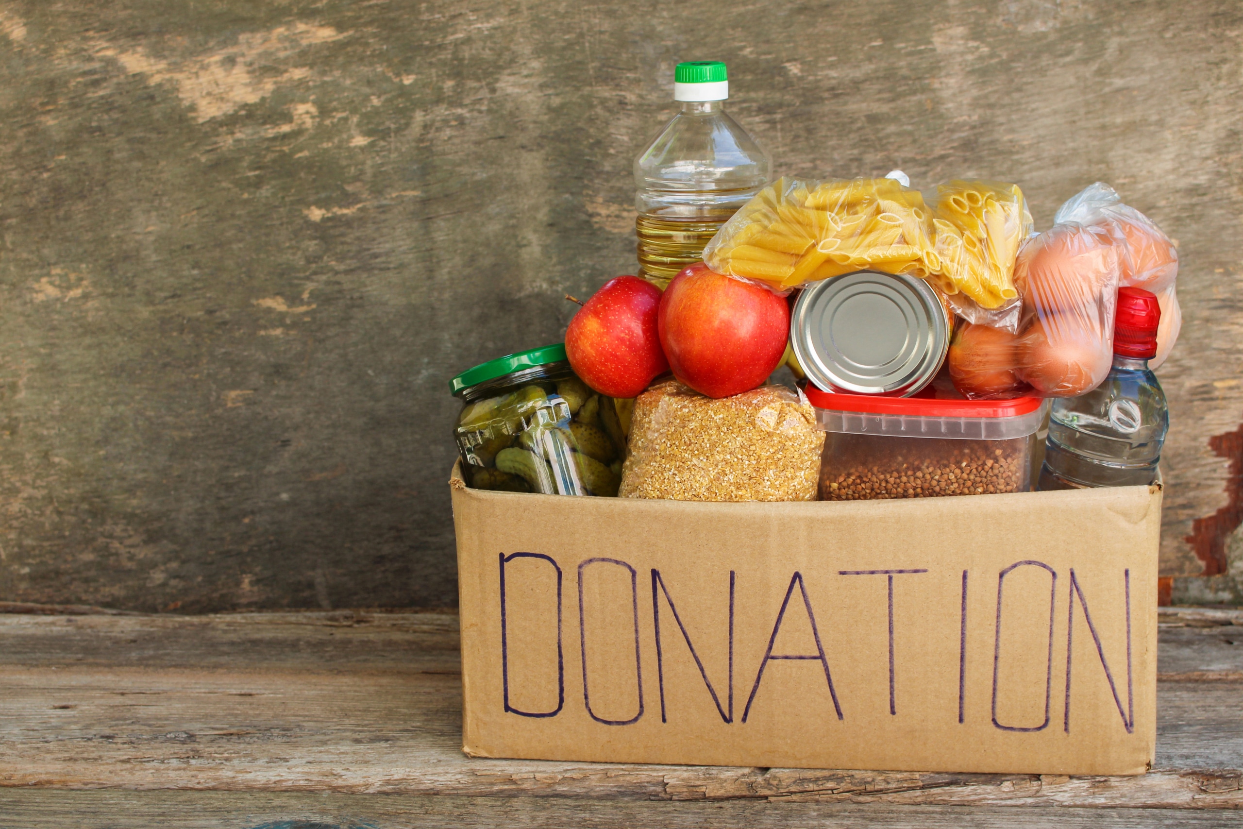 A cardboard box labeled DONATION is filled with various food items, including pasta, canned goods, apples, a bottle of oil, pickles in a jar, and eggs. The box is placed on a wooden surface with a rustic background.