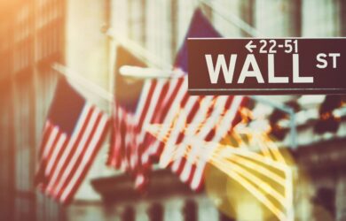Blurred image of several American flags in front of a building, with a prominent Wall Street sign in focus. Warm sunlight creates a soft glow, lending a historic and financial atmosphere.