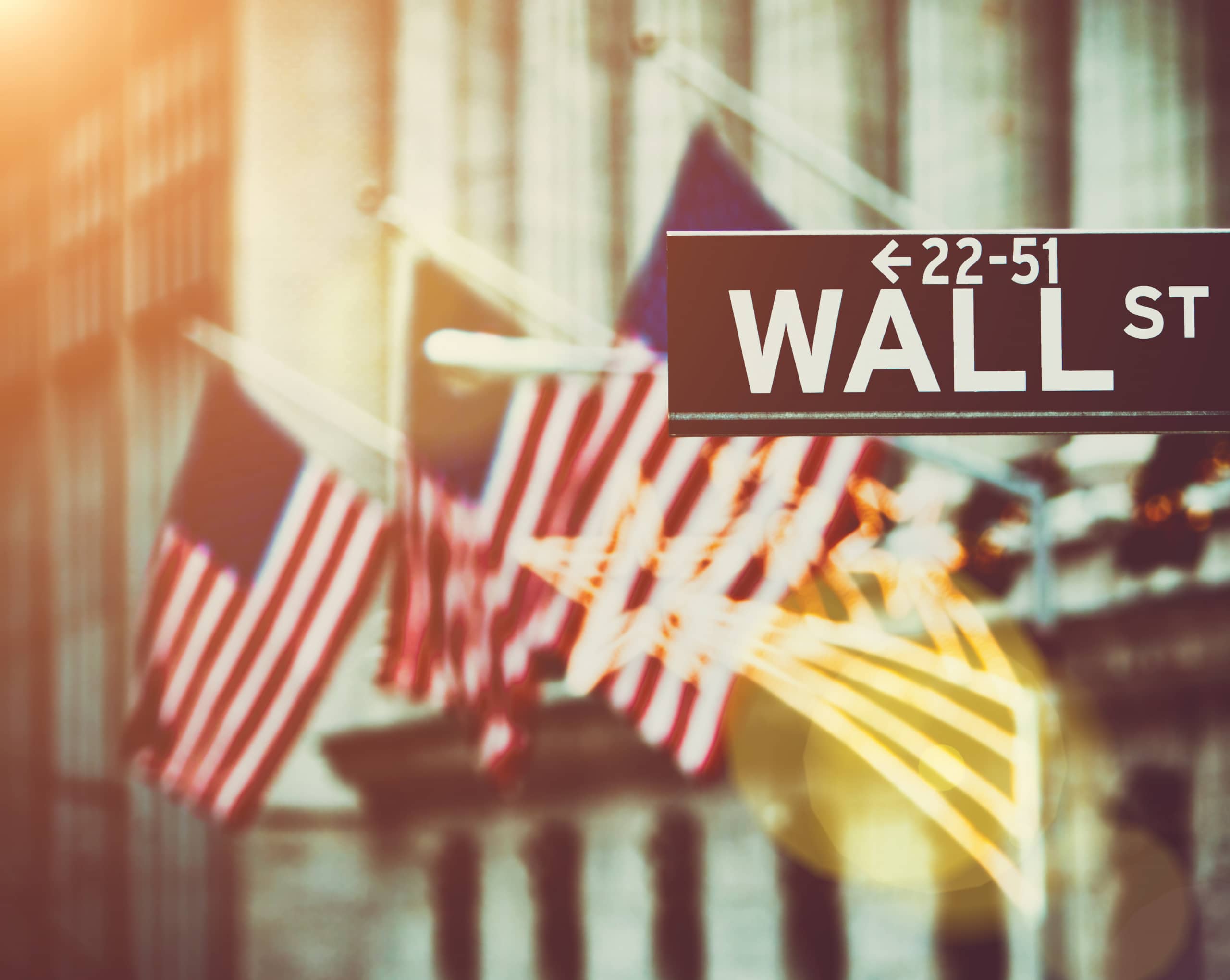 Blurred image of several American flags in front of a building, with a prominent Wall Street sign in focus. Warm sunlight creates a soft glow, lending a historic and financial atmosphere.