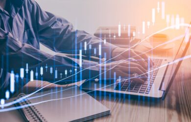 Person typing on a laptop with a digital stock market graph overlay. The graph features candlestick charts and rising lines, symbolizing financial analysis or trading. A notebook and a coffee cup are on the desk.