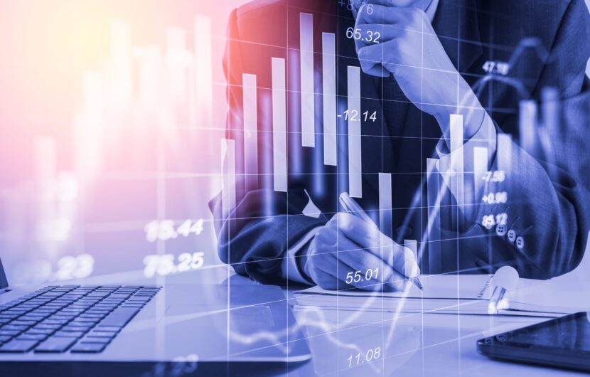 A person in a suit is writing on a paper at a desk with a laptop, surrounded by digital charts and graphs. The overlay of financial data suggests business analysis or stock market activities. The image has a blue and pink color tone.