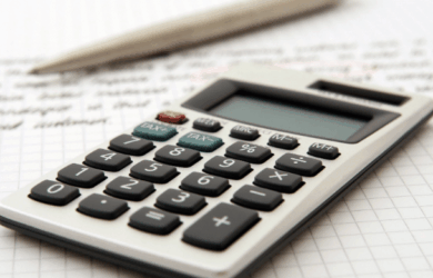 A calculator rests on grid paper with handwritten notes visible. A pen is placed next to the calculator, suggesting a work or study environment focused on calculations or financial tasks.
