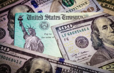 A close-up of a United States Treasury check is surrounded by various denominations of U.S. currency, including several one hundred dollar bills featuring Benjamin Franklin. The check displays the Statue of Liberty.
