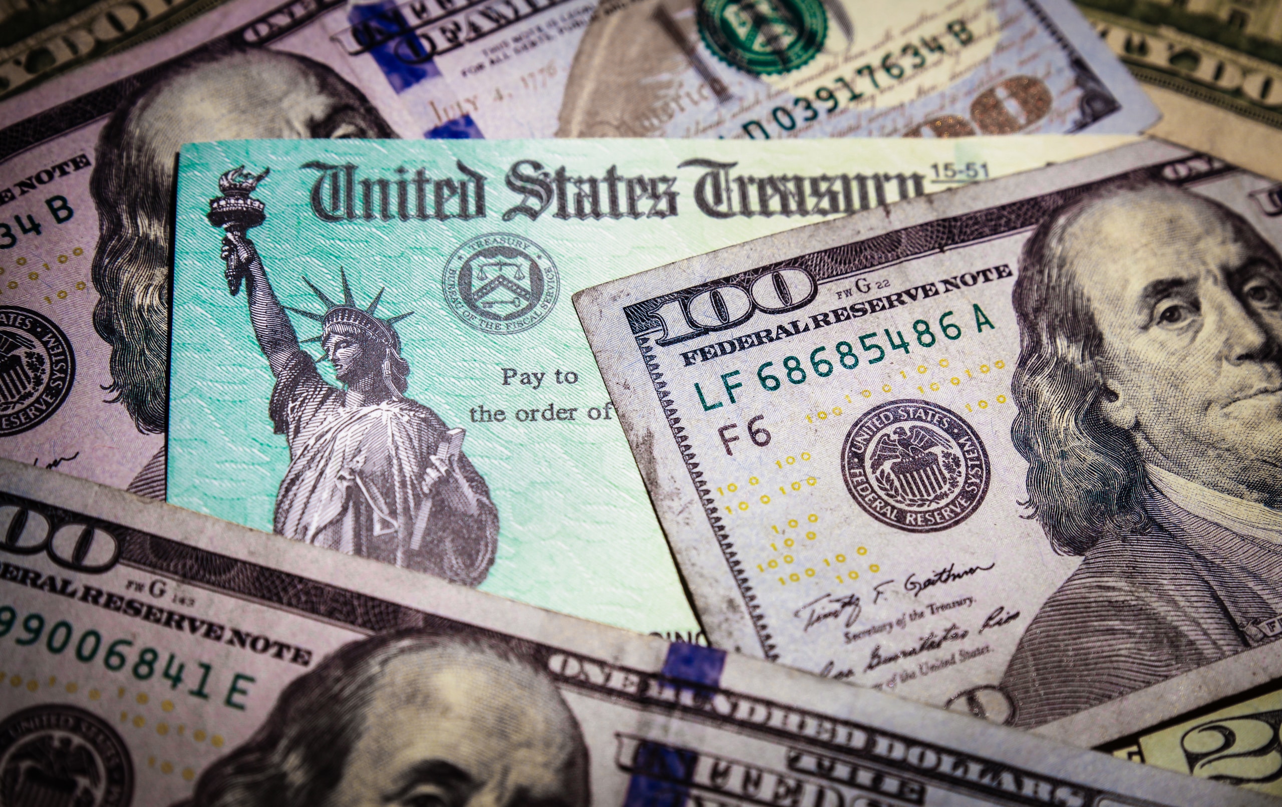 A close-up of a United States Treasury check is surrounded by various denominations of U.S. currency, including several one hundred dollar bills featuring Benjamin Franklin. The check displays the Statue of Liberty.