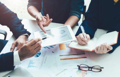 Three people collaborate around a table with financial documents and charts. One holds a tablet displaying graphs, another takes notes in a notebook, and a pair of glasses rests on the table. The scene conveys a business meeting atmosphere.