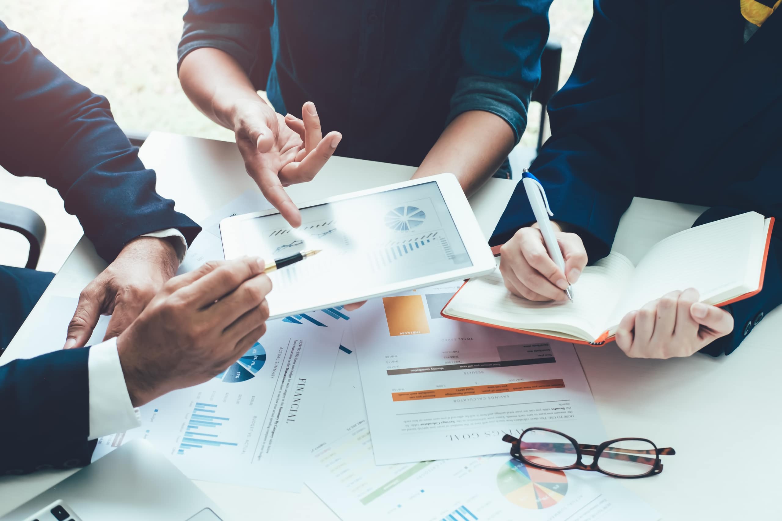 Three people collaborate around a table with financial documents and charts. One holds a tablet displaying graphs, another takes notes in a notebook, and a pair of glasses rests on the table. The scene conveys a business meeting atmosphere.