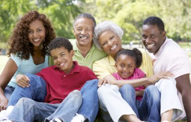 A joyful multi-generational family poses together outdoors. They are sitting on the grass, smiling brightly. The group includes grandparents, parents, and children, creating a warm and loving ambiance in a sunny park setting.