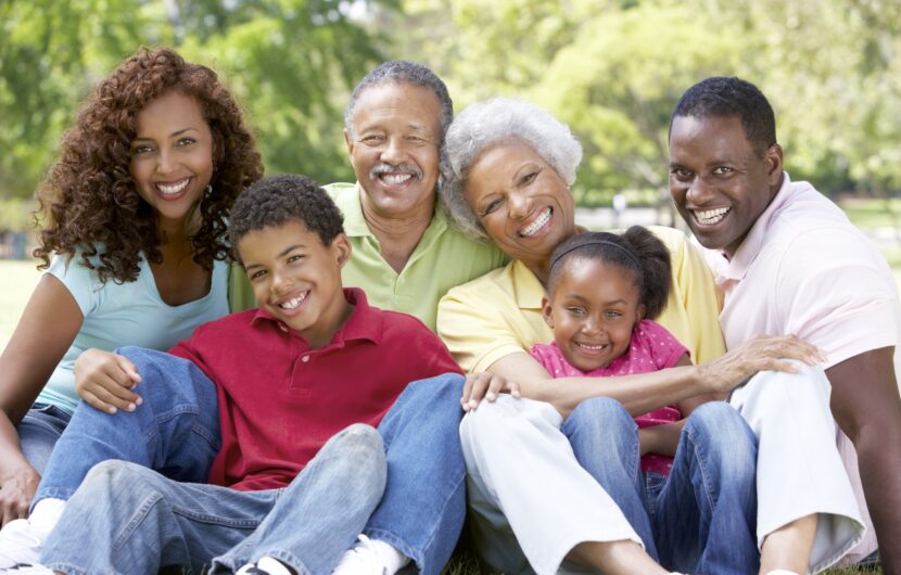 A joyful multi-generational family poses together outdoors. They are sitting on the grass, smiling brightly. The group includes grandparents, parents, and children, creating a warm and loving ambiance in a sunny park setting.