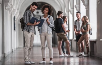 A group of students in a bright hallway. Two students in the foreground are looking at documents together, both wearing casual clothes and backpacks. Others in the background are engaged in conversation.