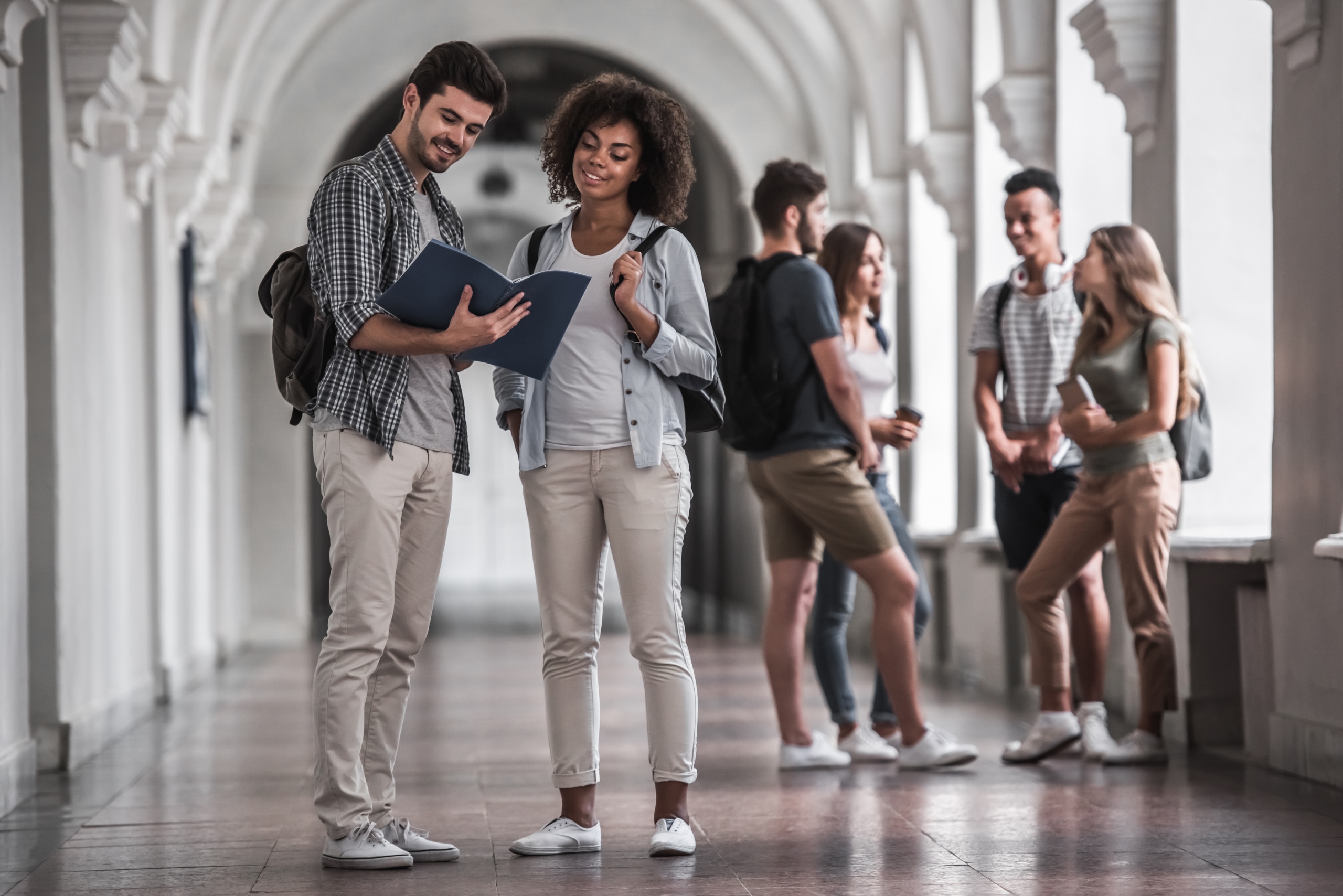 A group of students in a bright hallway. Two students in the foreground are looking at documents together, both wearing casual clothes and backpacks. Others in the background are engaged in conversation.
