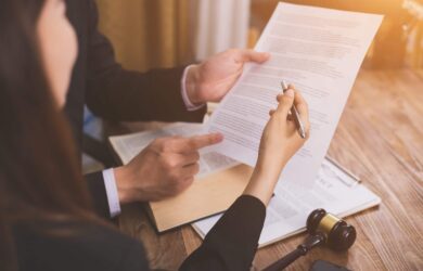 A person in a suit holds a document, while another with a pen reviews it. A wooden gavel rests on the table, suggesting a legal or formal setting on a wooden desk.