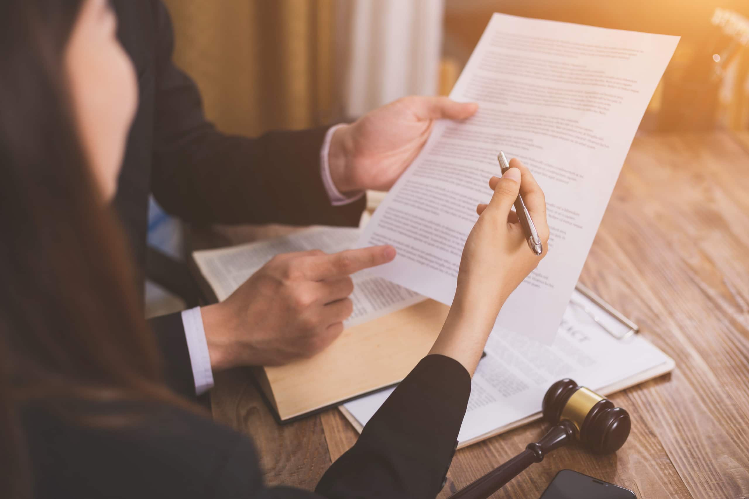 A person in a suit holds a document, while another with a pen reviews it. A wooden gavel rests on the table, suggesting a legal or formal setting on a wooden desk.