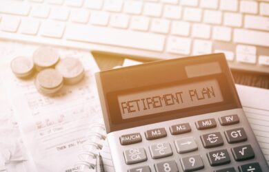 A calculator with RETIREMENT PLAN on the display is placed on sheets of paper, alongside a stack of coins. A computer keyboard is visible in the background. The scene is lit with warm lighting, suggesting financial planning or budgeting.