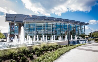 A modern glass building with a sign reading Savannah Convention Center. In front, a series of fountains and landscaped greenery. The sky is partly cloudy, and the sunlight highlights the architectural features.