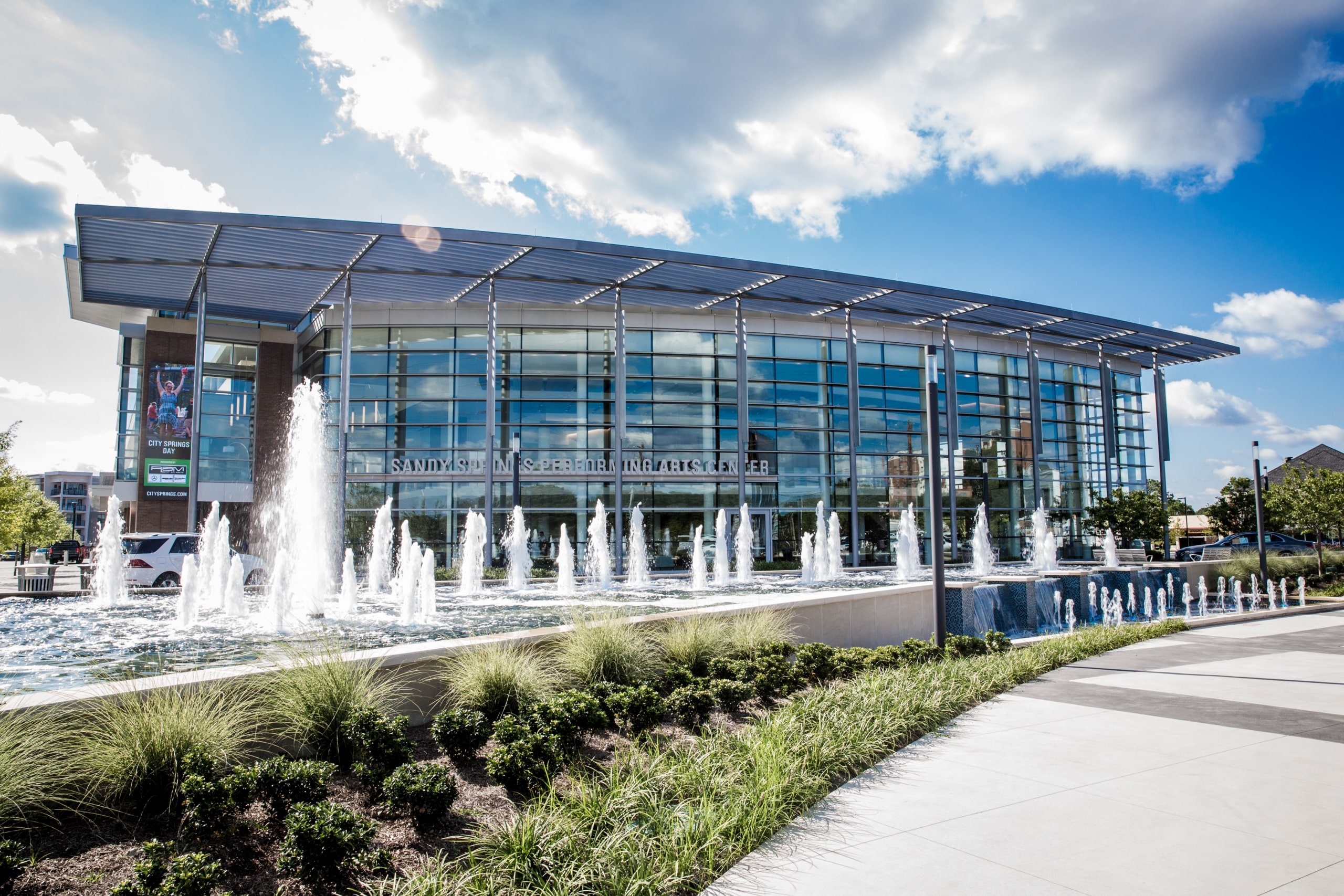 A modern glass building with a sign reading Savannah Convention Center. In front, a series of fountains and landscaped greenery. The sky is partly cloudy, and the sunlight highlights the architectural features.