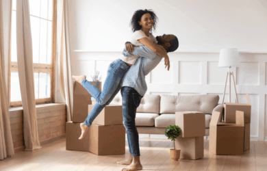 A joyful couple embraces in a bright, unfurnished living room with large windows. Cardboard boxes and a small potted plant are scattered on the hardwood floor, suggesting they are moving in. The woman is being lifted off her feet by the man.