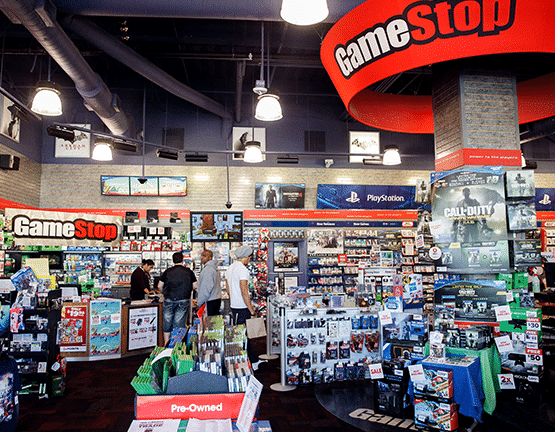 A GameStop store interior featuring various video game merchandise, including game cases, controllers, and posters. Several people are browsing the shelves, with prominent signage and logos visible, creating a colorful and vibrant gaming atmosphere.