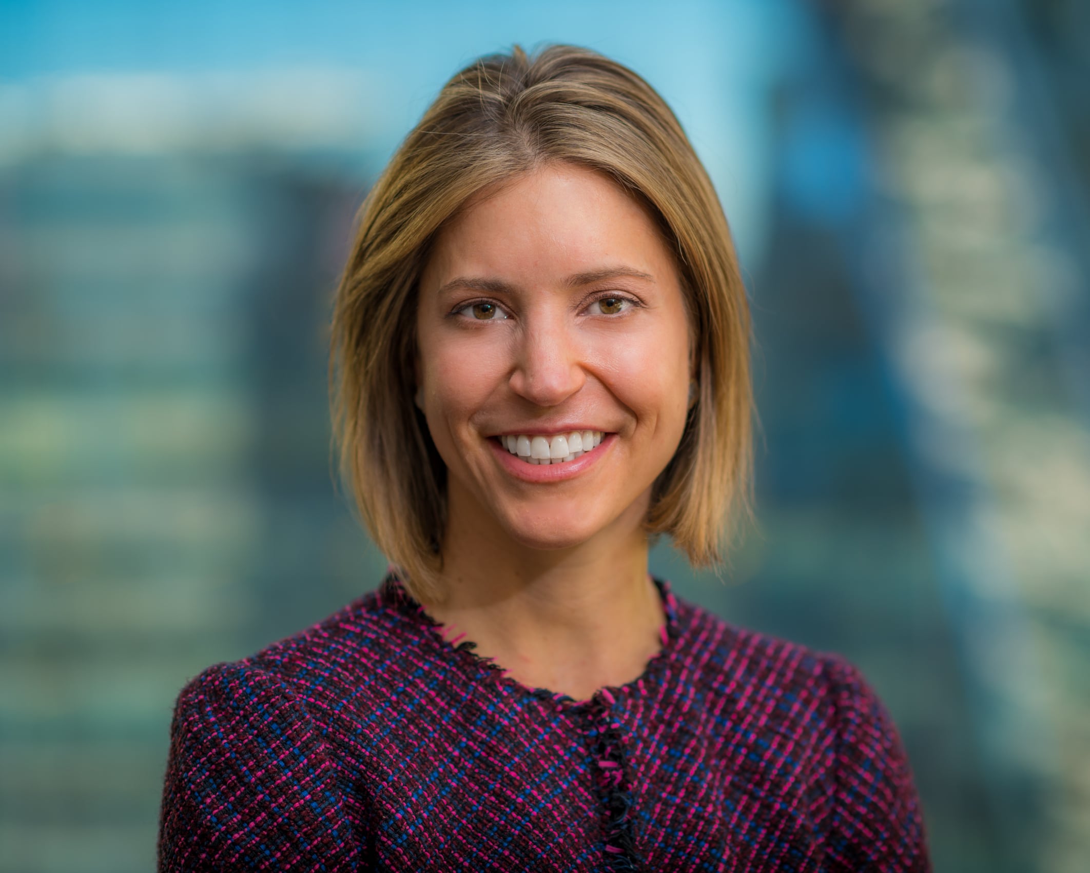 A smiling person with short, light brown hair is wearing a textured, dark-colored blazer. The background is blurred, featuring blue and green hues.