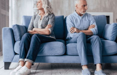 An elderly couple sits on a blue sofa, turned away from each other with crossed arms, appearing upset. The woman wears a gray sweater and white sneakers, and the man wears a striped shirt and blue shoes. The room has a soft, muted color palette.