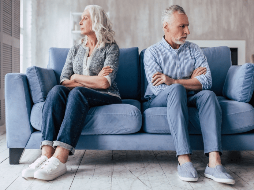 An elderly couple sits on a blue sofa, turned away from each other with crossed arms, appearing upset. The woman wears a gray sweater and white sneakers, and the man wears a striped shirt and blue shoes. The room has a soft, muted color palette.