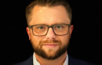 A man with short hair and a beard, wearing glasses and a dark suit with a light striped shirt, stands against a black background.