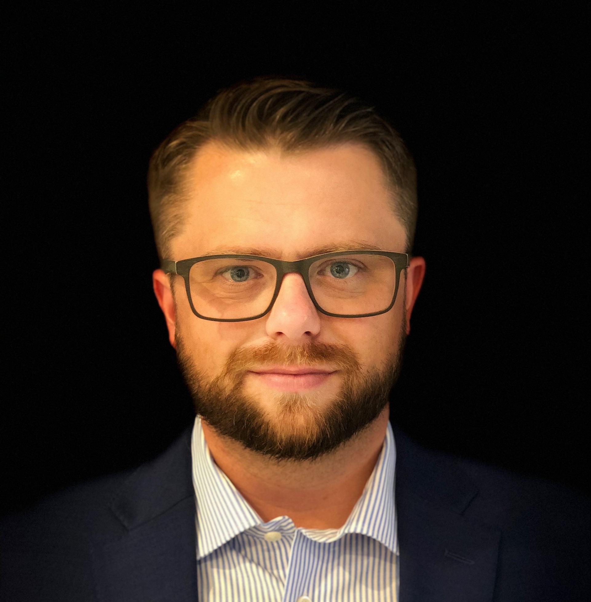 A man with short hair and a beard, wearing glasses and a dark suit with a light striped shirt, stands against a black background.