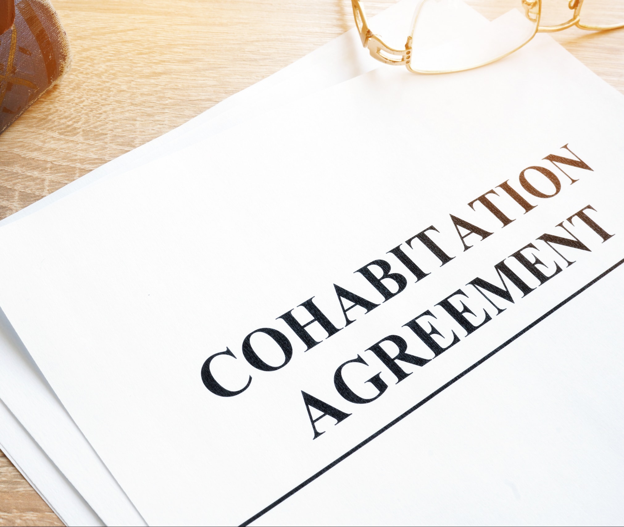A document titled Cohabitation Agreement rests on a wooden surface. Nearby, a pair of glasses is partially visible, suggesting review or signing. The scene is well-lit, emphasizing the importance of the paperwork.