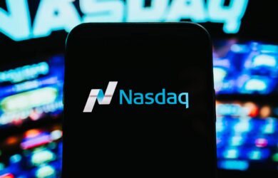 A smartphone displays the Nasdaq logo in white and blue against a blurred background of the Nasdaq sign in neon lights. The image suggests a focus on financial markets or stock exchange.