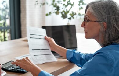 A woman with gray hair and glasses is sitting at a table, holding a document in one hand and using a calculator with the other. A laptop and potted plant are on the table, and soft light filters through a window in the background.