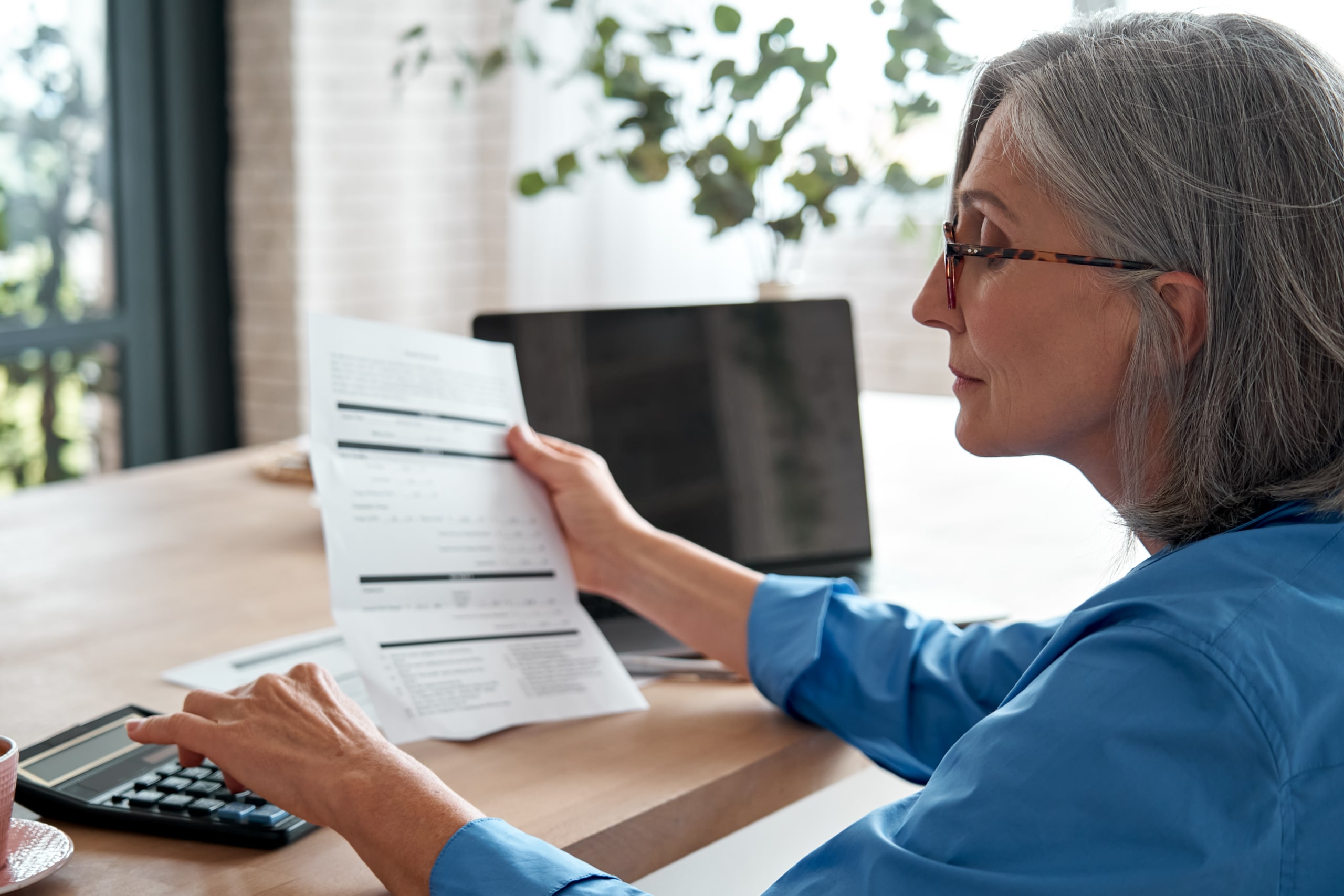 A woman with gray hair and glasses is sitting at a table, holding a document in one hand and using a calculator with the other. A laptop and potted plant are on the table, and soft light filters through a window in the background.