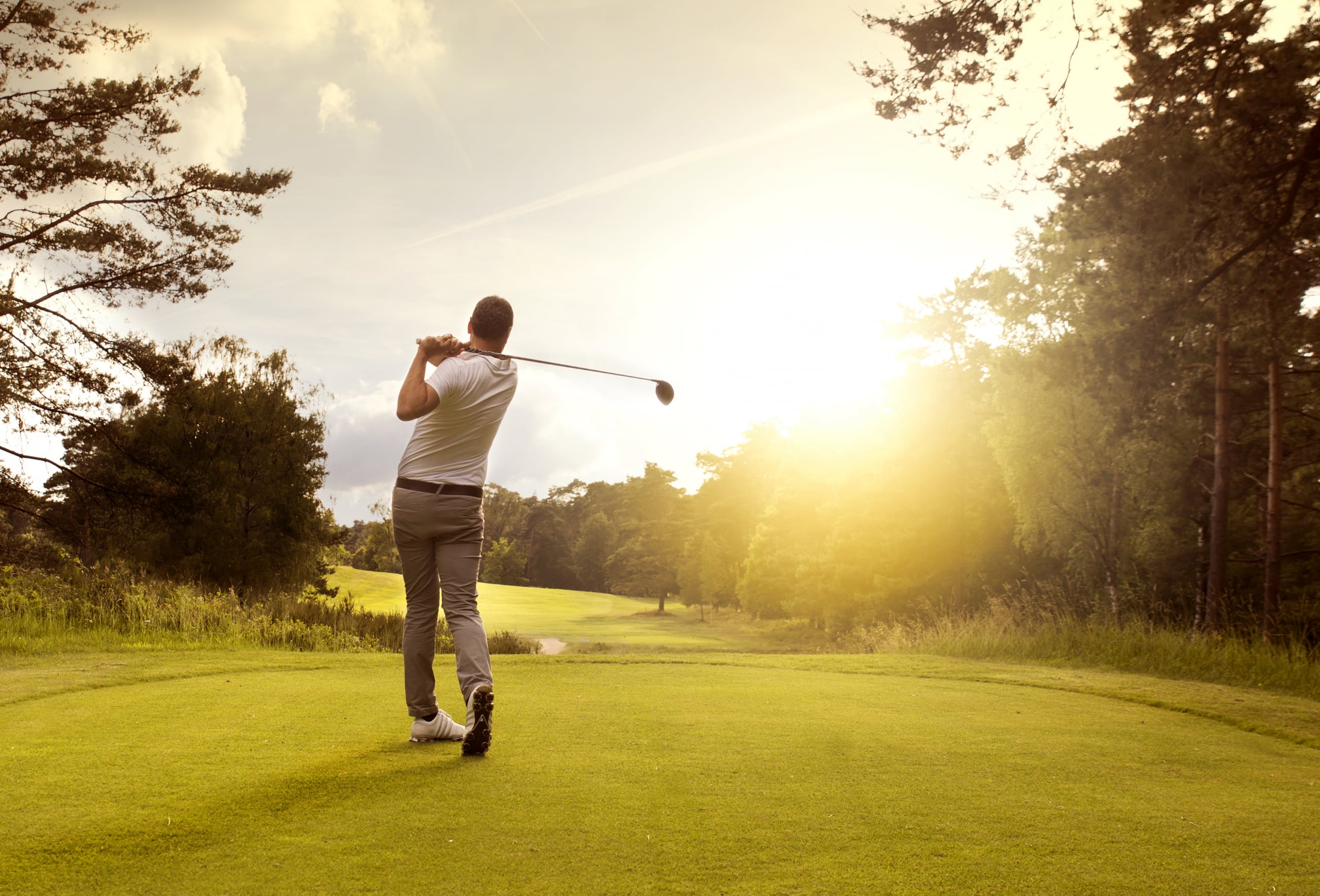 A golfer wearing a white shirt and gray pants swings a club on a sunny golf course. The sun is setting in the background, casting a warm glow over the green landscape bordered by trees.