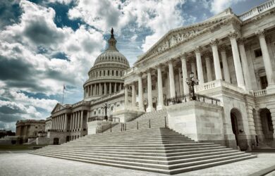 A dramatic view of the United States Capitol building under a cloudy sky. The image features the grand staircase leading up to the impressive façade, with its iconic dome and classic architectural columns.