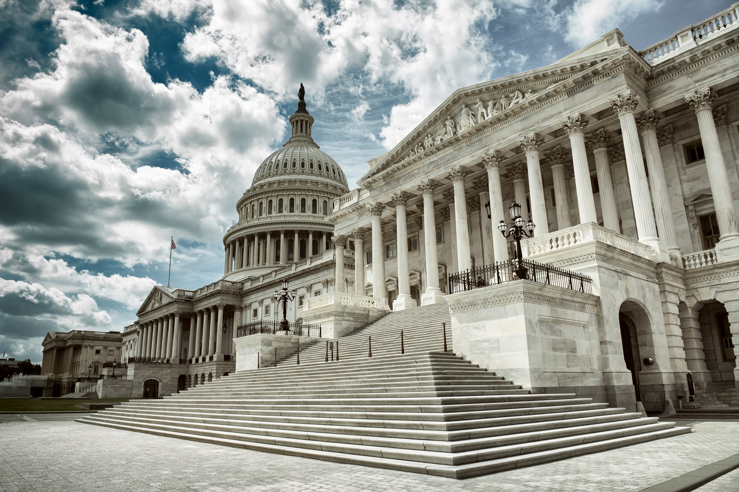 A dramatic view of the United States Capitol building under a cloudy sky. The image features the grand staircase leading up to the impressive façade, with its iconic dome and classic architectural columns.