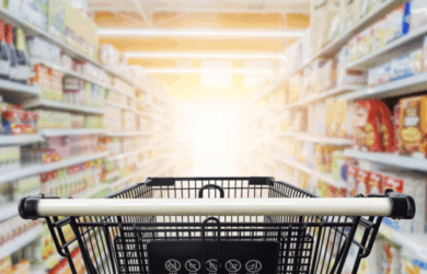 A shopping cart is centered in the aisle of a brightly lit grocery store. Shelves on either side are stocked with various products, and the far end is illuminated by a soft, glowing light.
