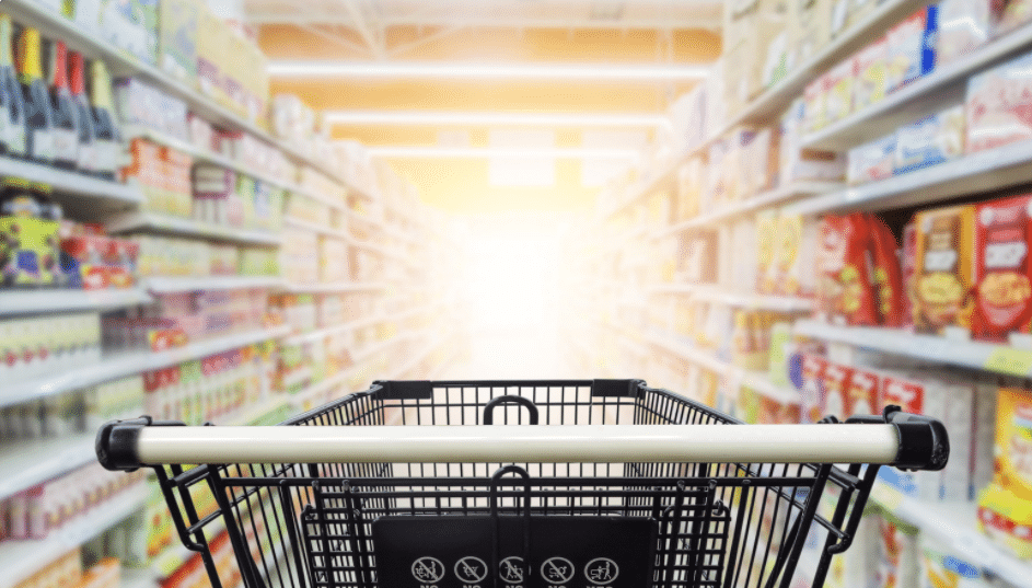 A shopping cart is centered in the aisle of a brightly lit grocery store. Shelves on either side are stocked with various products, and the far end is illuminated by a soft, glowing light.