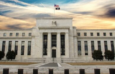 The image shows the exterior of the Federal Reserve Building, a large rectangular structure with a central entrance, tall columns, and an American flag atop. The sky is partly cloudy with blue, orange, and yellow hues.